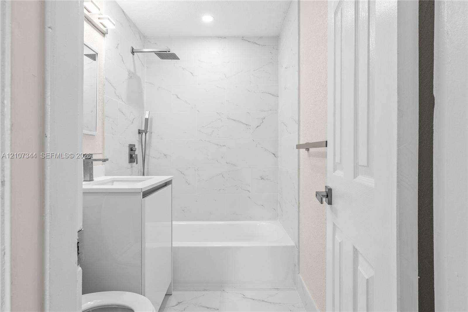 Full-height stone-finish wall tile, white vanity cabinet, rectangular undermount sink, rainfall showerhead with handheld sprayer, and recessed lighting