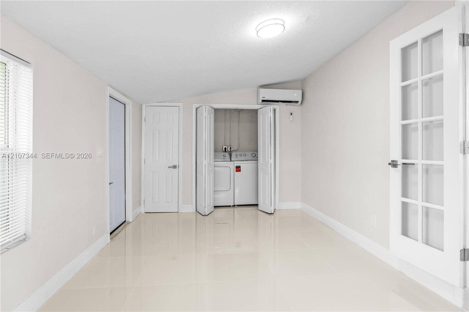 Interior space featuring light-toned tile flooring, recessed lighting, a dedicated closet with bi-fold doors containing laundry appliances, wall-mounted air conditioning unit, and a French door with glass panels