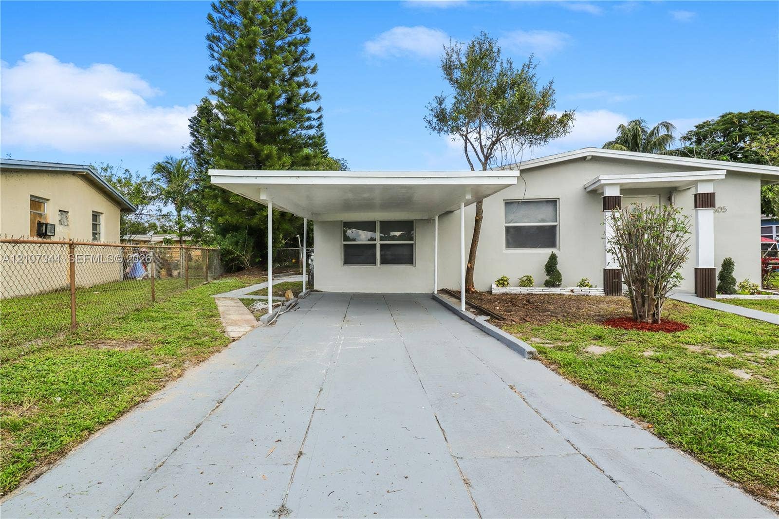 Single-story exterior featuring a painted concrete facade, attached carport with support pillars, concrete driveway, and covered front entryway with decorative columns