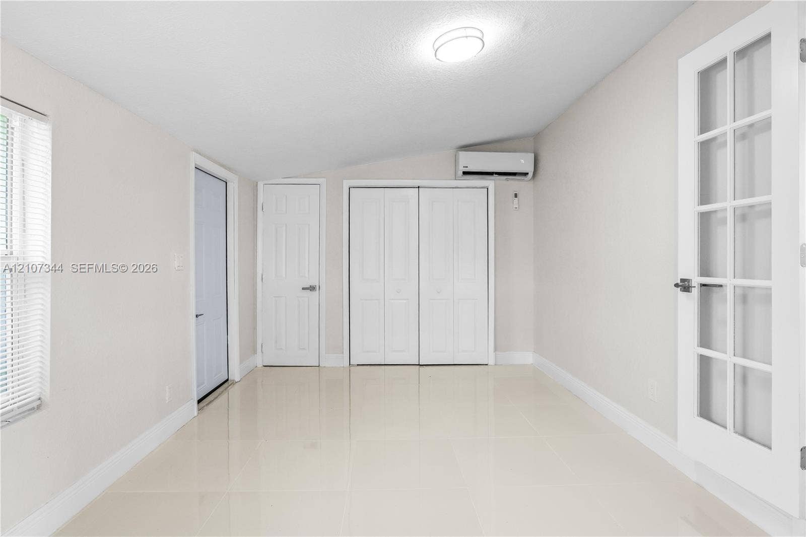 Neutral-toned interior featuring large-format tile flooring, double-door closet cabinetry, wall-mounted ductless climate control unit, and a French-style door with glass paneling