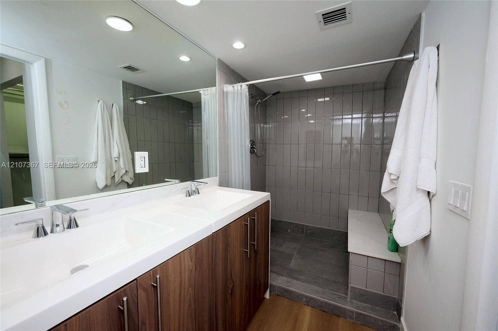 2851 NE 183rd Street, #1617E, Aventura, FL 33160 Double vanity with rectangular integrated sinks, wood-finish cabinetry, full-width mirror, and walk-in shower featuring a tiled bench seat