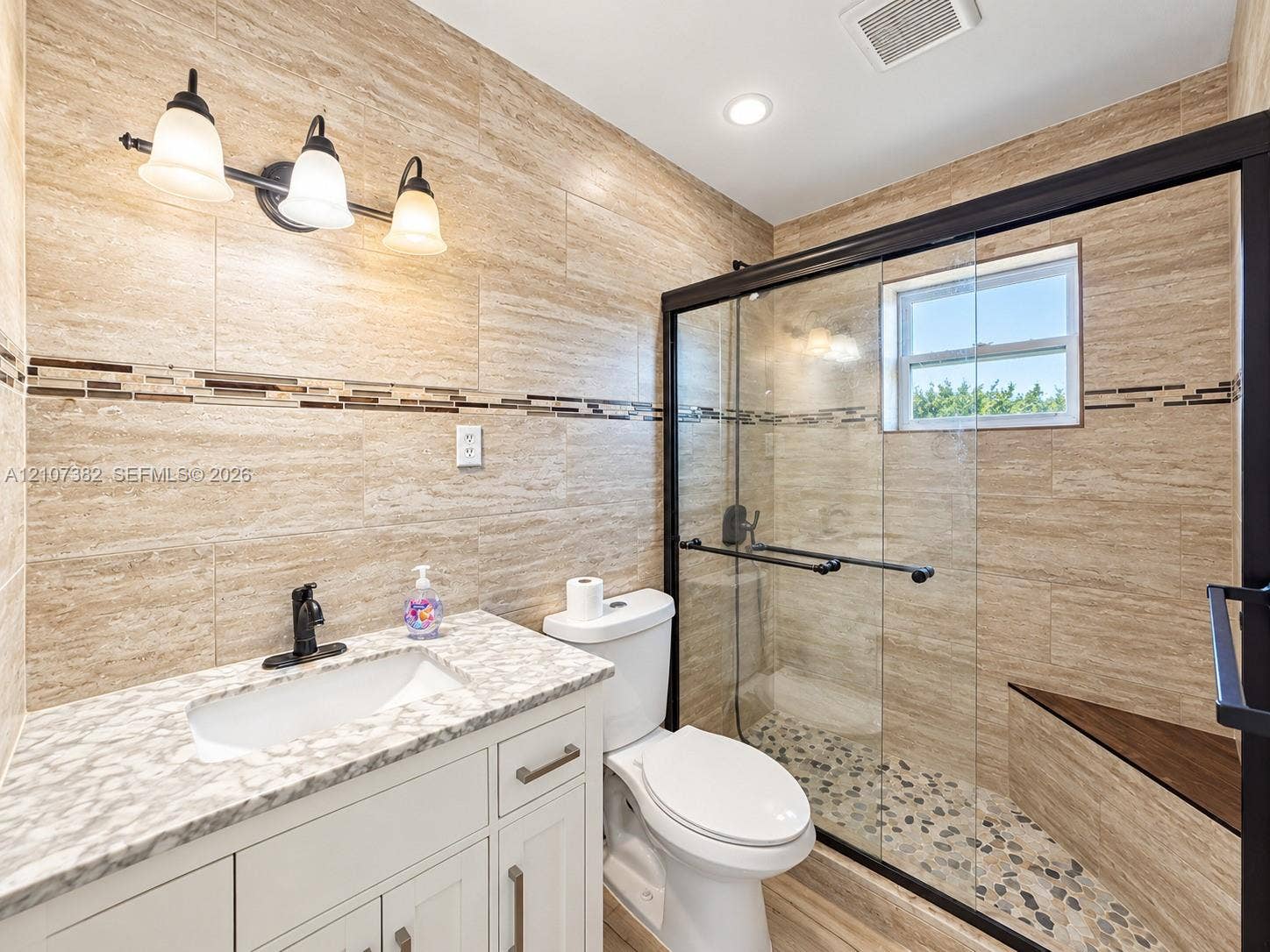 859 Caroline Circle, Palm Beach, FL 33413 Full bathroom featuring floor-to-ceiling stone-look wall tile, vanity with a patterned countertop, glass-enclosed shower with pebble-stone flooring, and a built-in shower bench