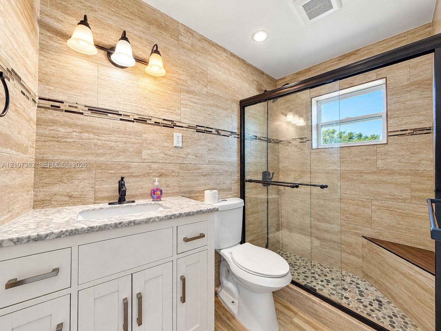 859 Caroline Circle, Palm Beach, FL 33413 Bathroom featuring large-format wall tiling with decorative mosaic inlay, stone-composite countertop with integrated sink, shaker-style cabinetry, and a walk-in shower with pebble-stone flooring and a built-in bench