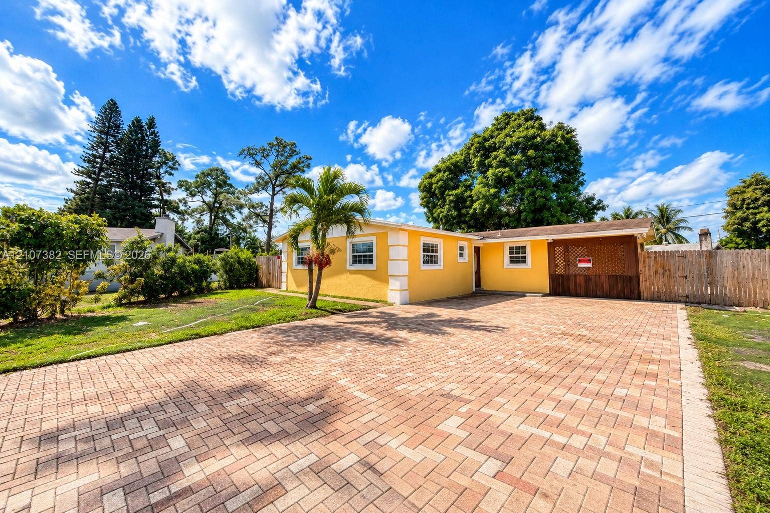 859 Caroline Circle, Palm Beach, FL 33413 Exterior facade featuring a yellow stucco finish, expansive brick-paver driveway, integrated carport, and wood-latched privacy fencing