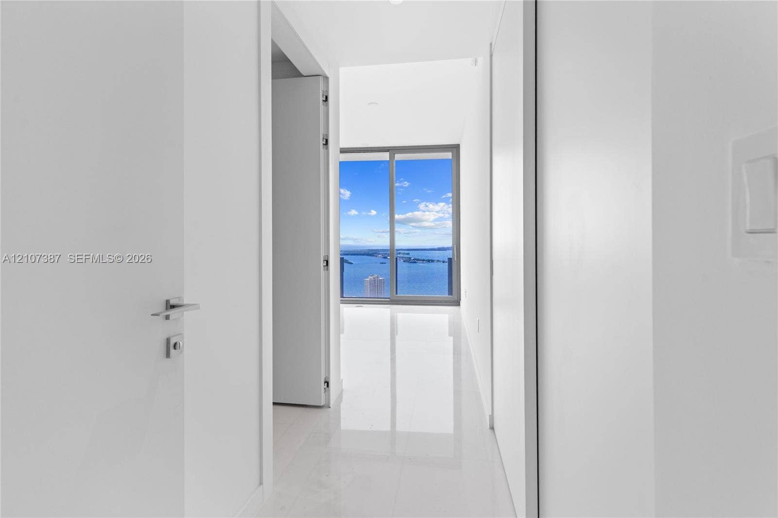 Hallway featuring polished white tile flooring, sleek flat-panel doors with contemporary hardware, and floor-to-ceiling sliding glass doors highlighting a water backdrop