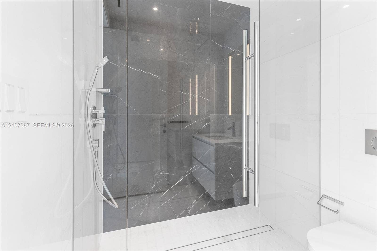Walk-in shower featuring large-format dark veined tile surround, frameless glass enclosure, handheld shower fixture, and linear drain
