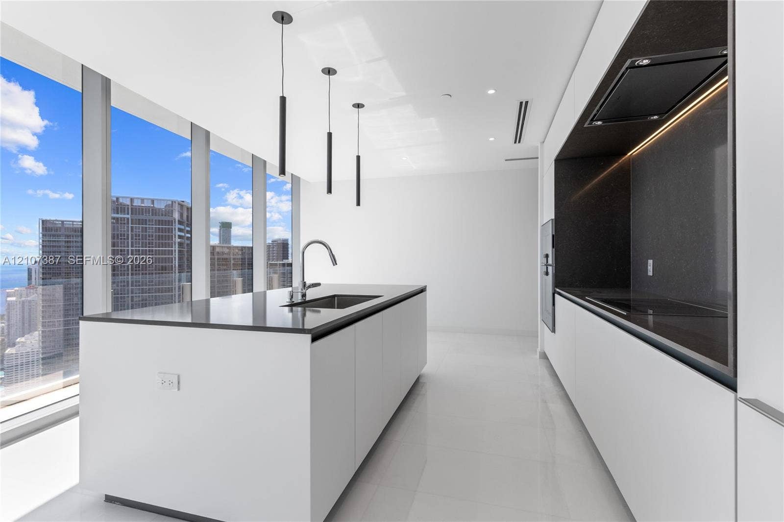 Kitchen featuring white flat-panel cabinetry, a central island with a contrasting stone-finish countertop, stainless steel undermount sink, floor-to-ceiling windows, and linear pendant lighting