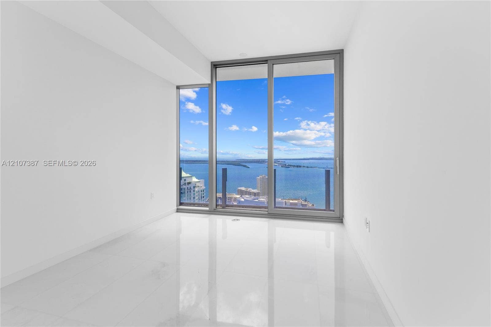 Floor-to-ceiling windows with sliding glass door access to a balcony, polished white stone-finish flooring, and expansive water scenery