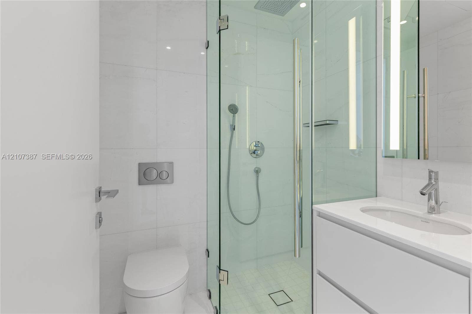 Bathroom featuring a glass-enclosed shower with a rainfall showerhead, wall-mounted dual-flush toilet, white vanity with an integrated sink, polished chrome fixtures, and large-format tile surround