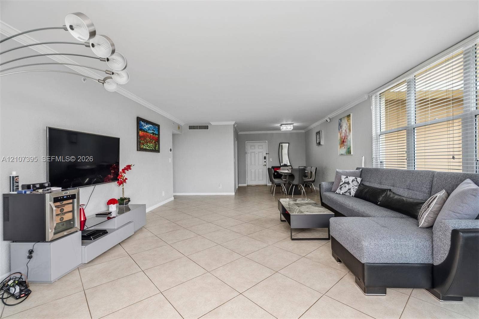 Open-concept interior featuring large-format diagonal tile flooring, crown molding, multi-arm arched floor lighting, and expansive windows with horizontal blinds