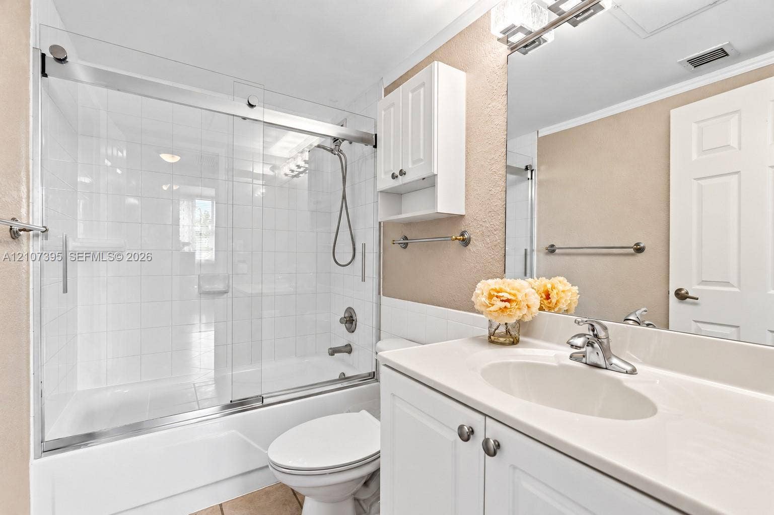 Bathroom featuring a white vanity cabinet with an integrated sink, polished chrome faucet, glass-enclosed shower and tub combination with tile surround, and a wall-mounted storage cabinet