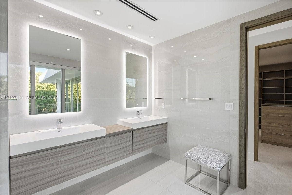 Floating double vanity with wood-finish cabinetry, integrated rectangular sinks, polished chrome fixtures, back-lit vanity mirrors, and floor-to-ceiling large-format tile wall cladding