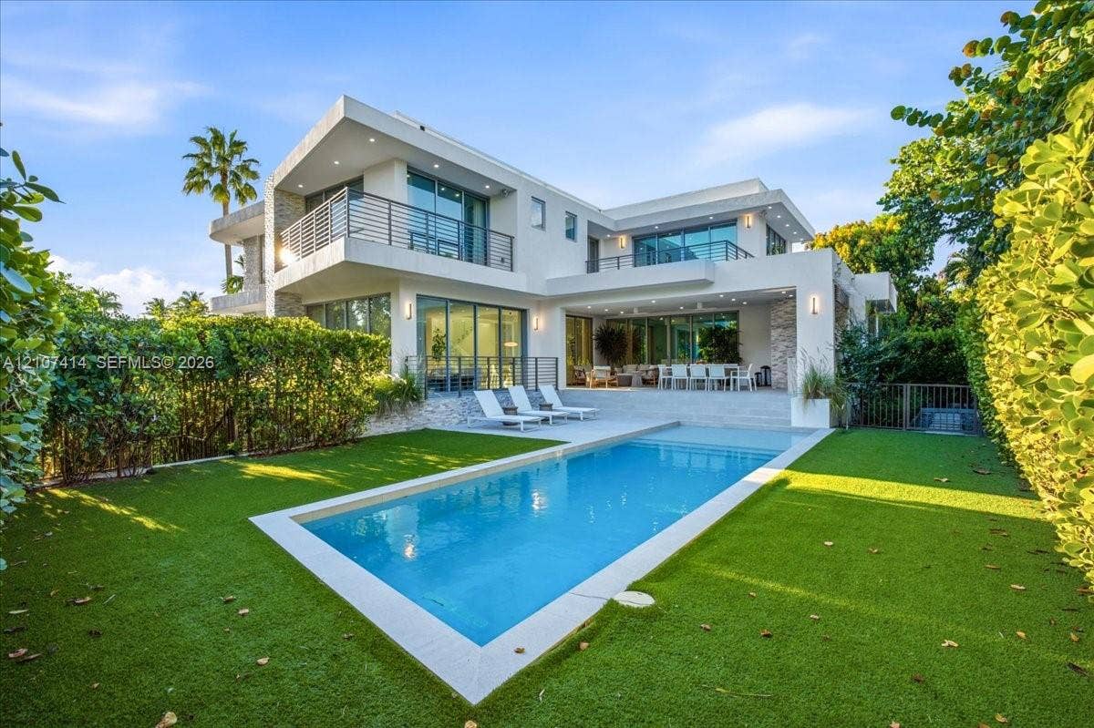 Modern two-story exterior featuring smooth stucco finish, stone accent pillars, expansive glass sliding doors, elevated balconies with metal railings, and a rectangular swimming pool surrounded by a manicured lawn