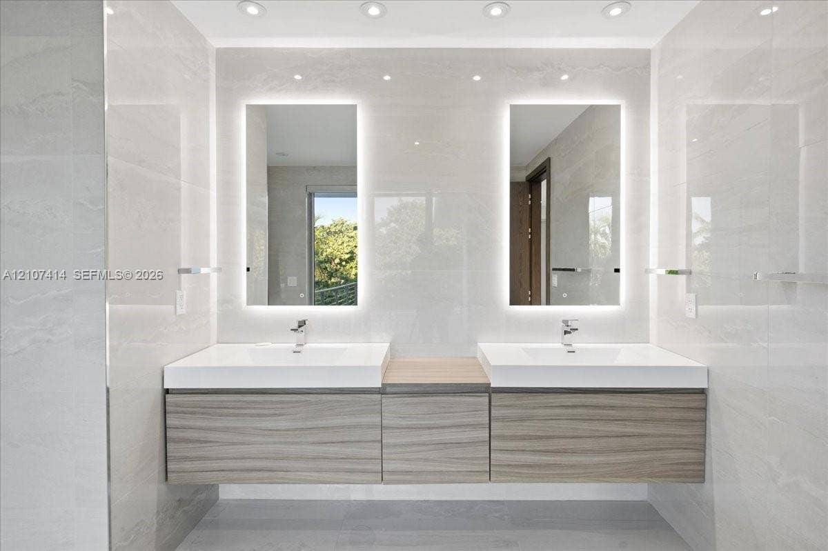 Floating dual vanity with wood-finish cabinetry, white integrated sinks, backlit rectangular mirrors, and recessed ceiling lighting, set against large-format tiled walls