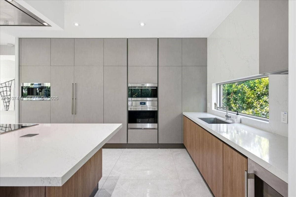 Modern kitchen featuring floor-to-ceiling flat-panel cabinetry, a large central island with white quartz countertops, integrated stainless steel appliances, wood-finish lower cabinetry, and a horizontal window backsplash