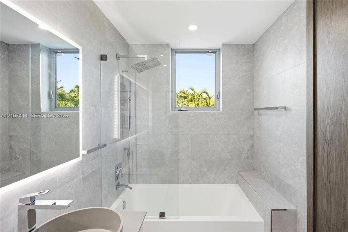 Bathroom featuring floor-to-ceiling stone-finish tile, an integrated soaking tub with a glass panel, a built-in stone ledge, and a backlit mirror