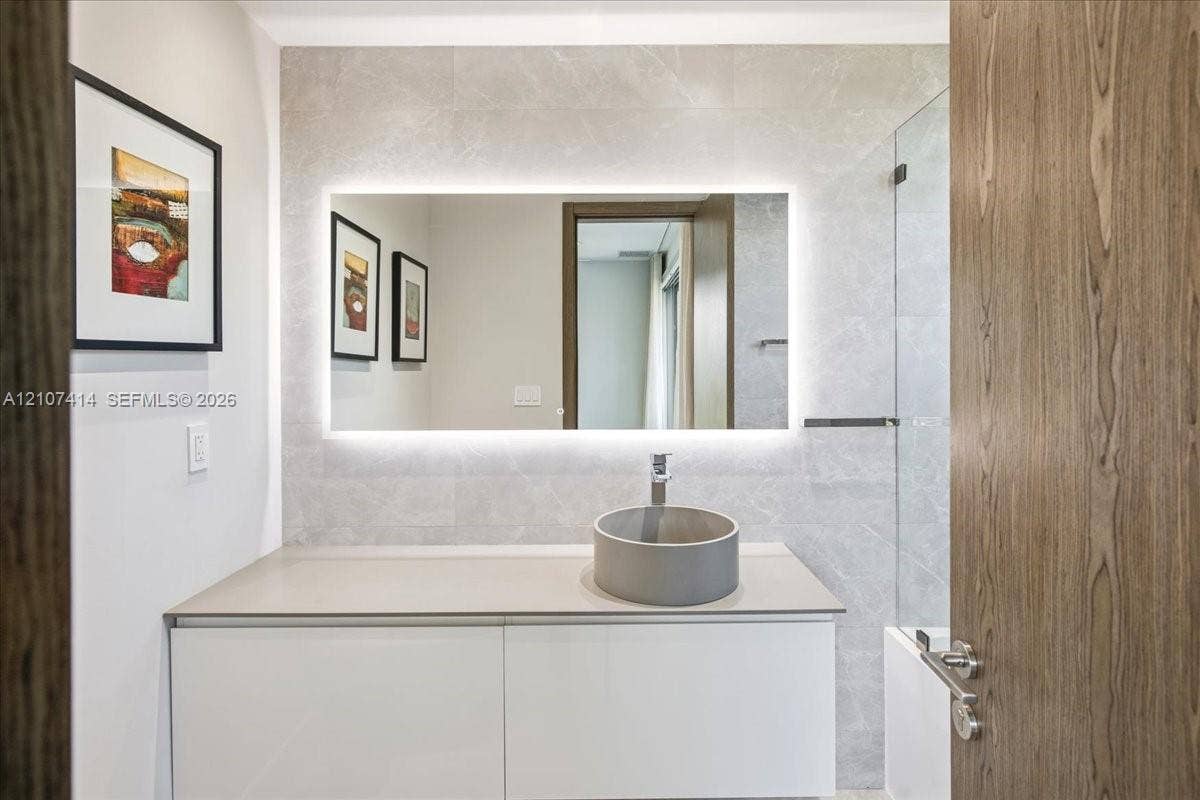White vanity with a solid-surface countertop, cylindrical vessel sink, LED-backlit mirror, and full-height stone-finish wall tiling