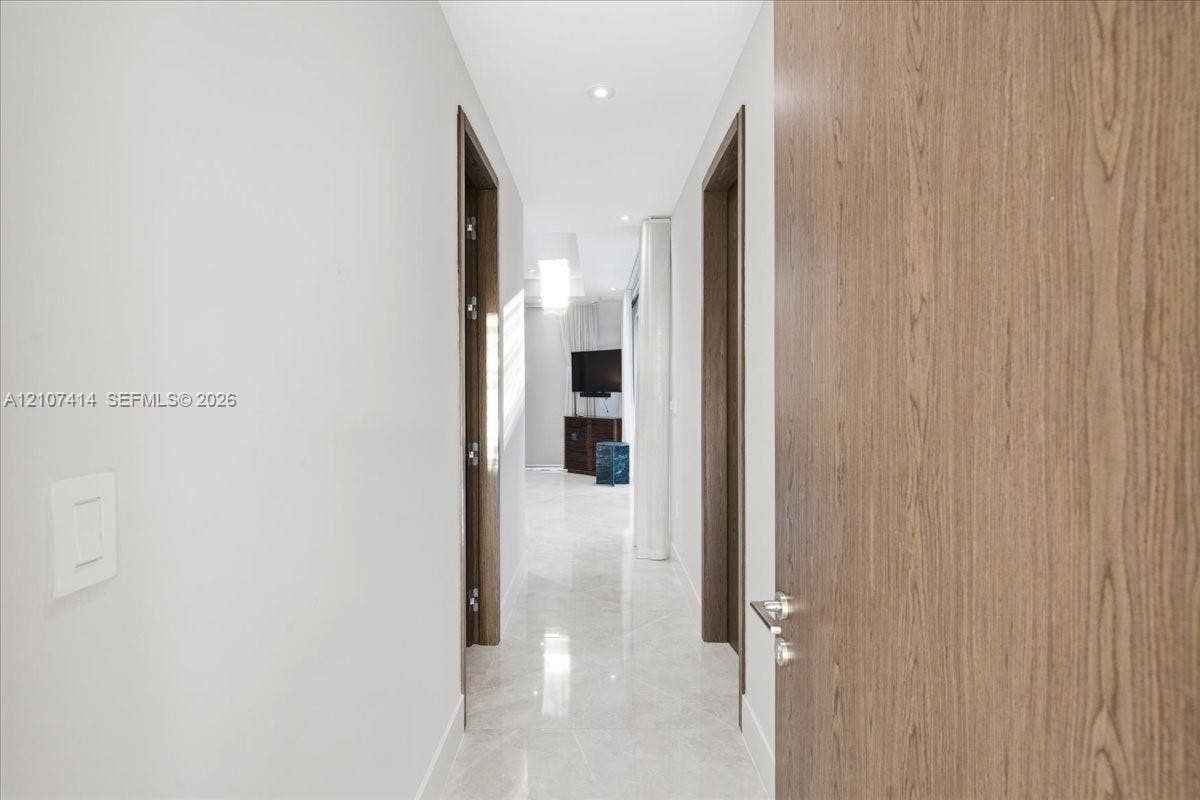 Hallway featuring large-format light-toned stone flooring, recessed ceiling lighting, and solid wood-finish doors with brushed-metal hardware