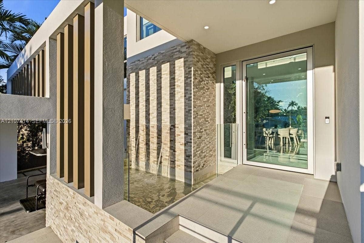 Modern entryway featuring a stacked stone facade, vertical architectural slats, an integrated water feature, recessed ceiling lighting, and a full-height glass-paneled entry door