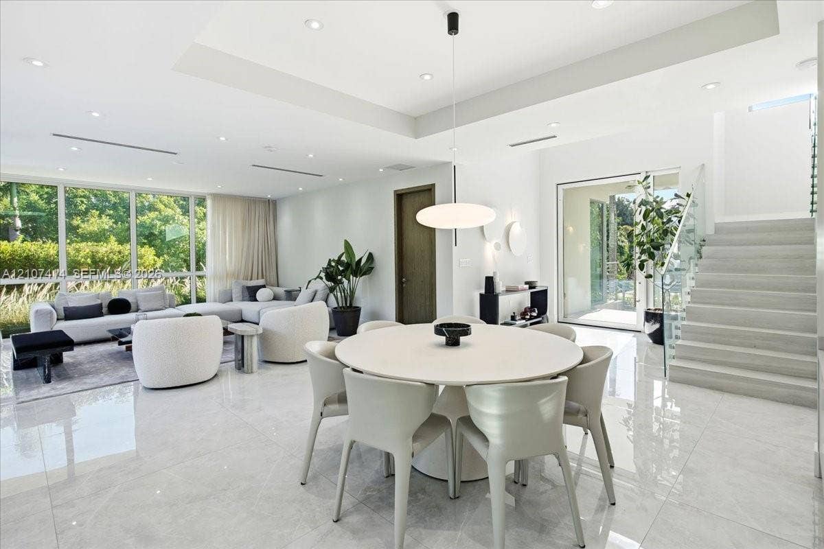 Open-concept interior featuring large-format polished stone flooring, tray ceilings with recessed lighting, floor-to-ceiling windows, and a staircase with a glass-panel railing