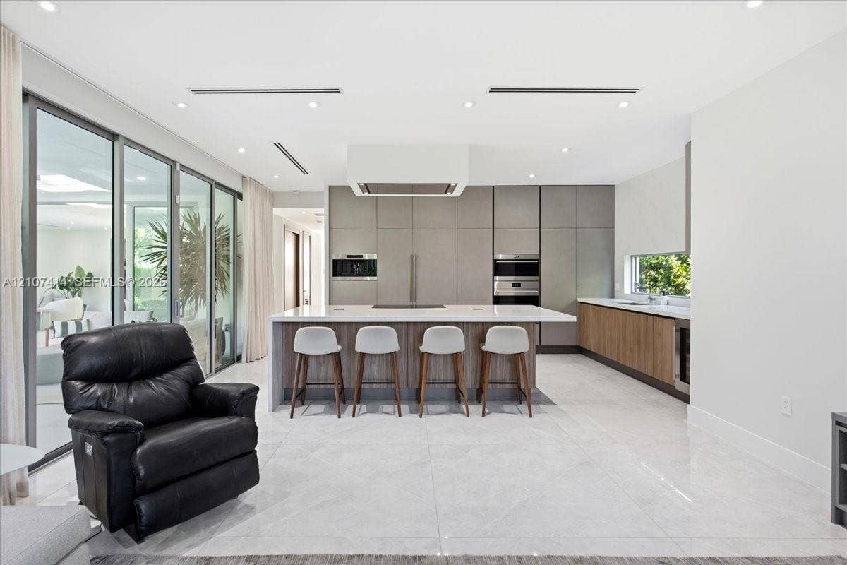 Chefs kitchen featuring full-height matte cabinetry, a waterfall-edge stone island, built-in stainless steel appliances, polished stone flooring, and expansive floor-to-ceiling glass sliding doors