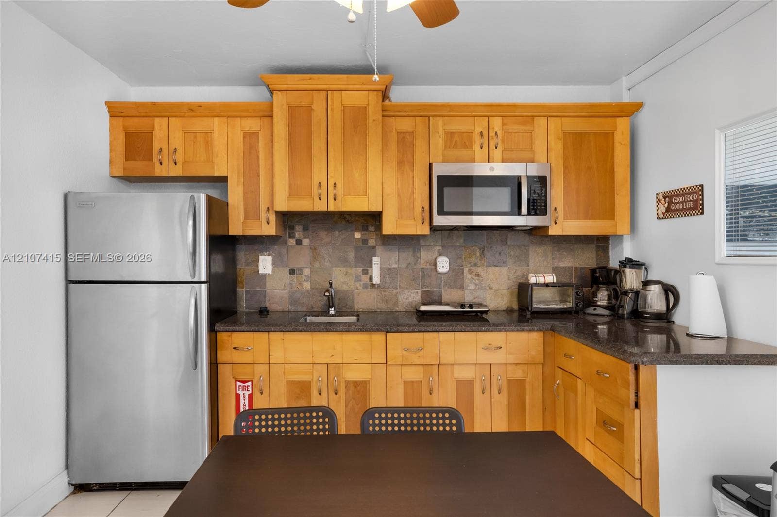 Kitchen featuring natural wood cabinetry, stone-tile backsplash, dark stone countertops, stainless steel refrigerator, and built-in microwave