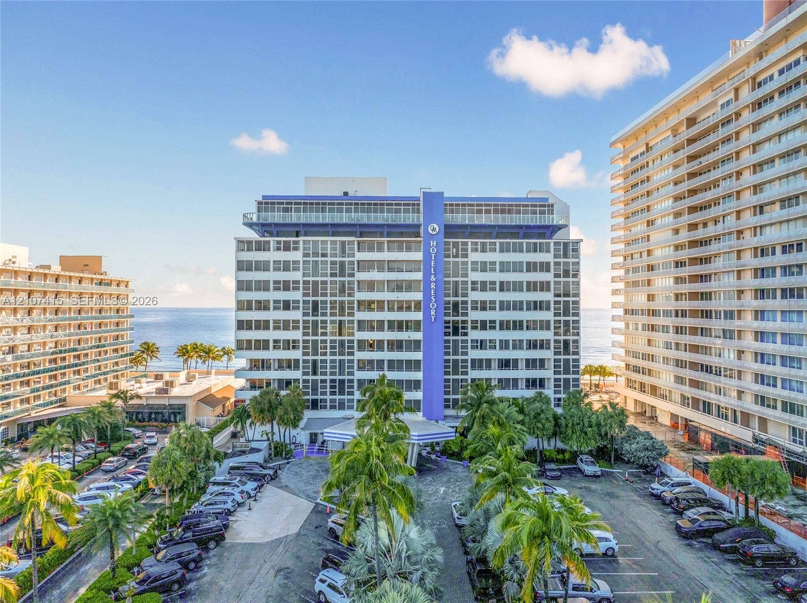 High-rise coastal building featuring multi-level balconies, expansive glass windows, and paved surface parking framed by mature palm trees