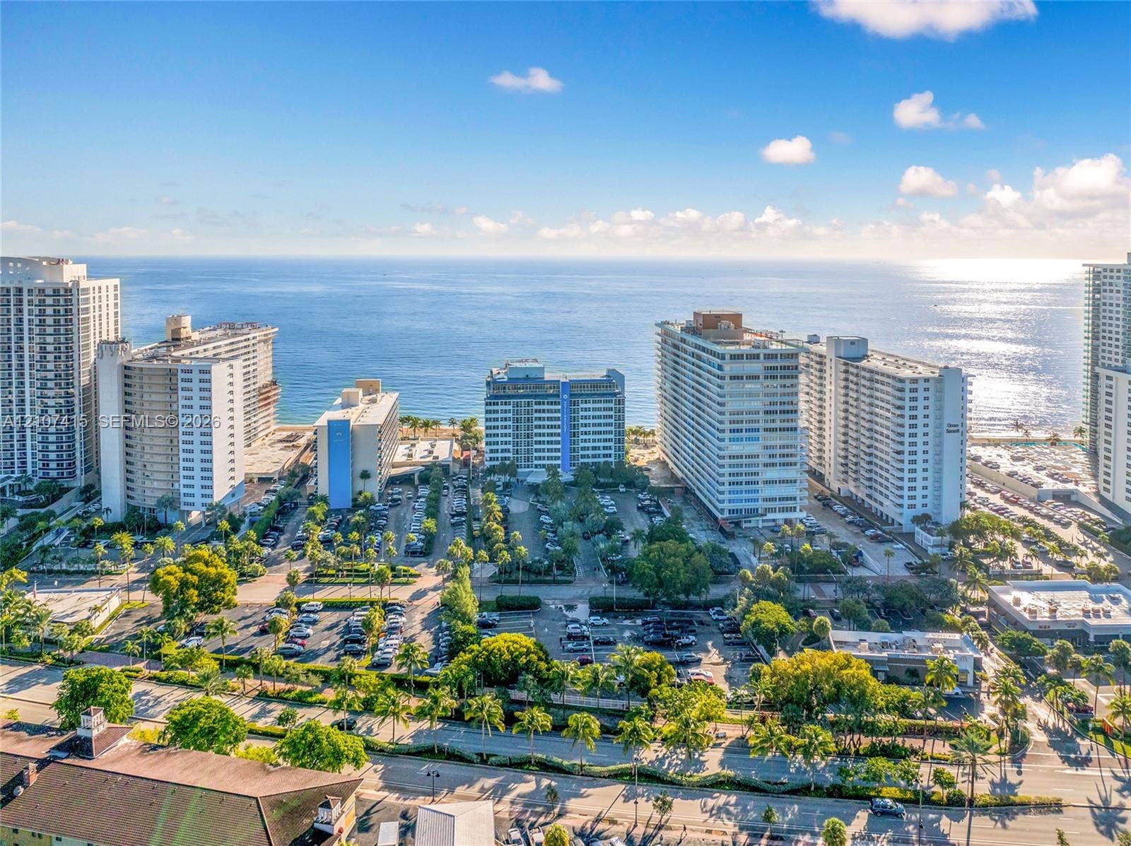 Aerial perspective featuring coastal proximity, high-rise architecture, mature canopy trees, and paved surface parking
