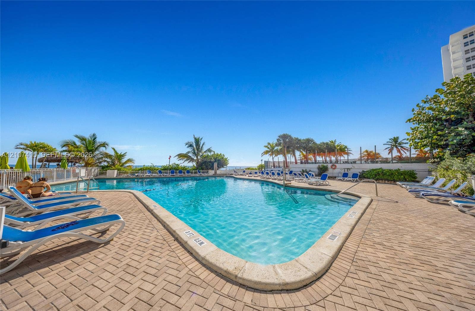 Swimming pool featuring a herringbone-patterned paver deck, metal perimeter fencing, integrated steps, and mature palm trees