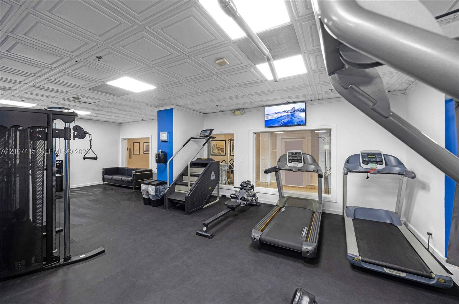 Fitness room featuring patterned drop ceiling tiles, recessed lighting, rubberized flooring, and large glass-paneled partitions