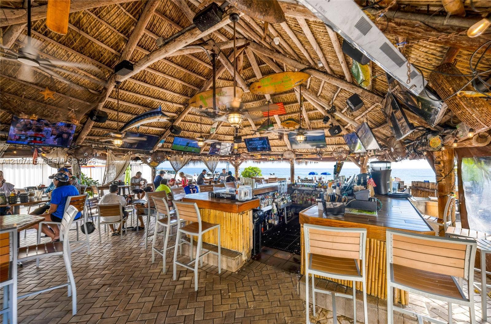 Open-air pavilion featuring a vaulted thatched roof, exposed timber framing, a central bar with bamboo-style paneling, and brick paver flooring