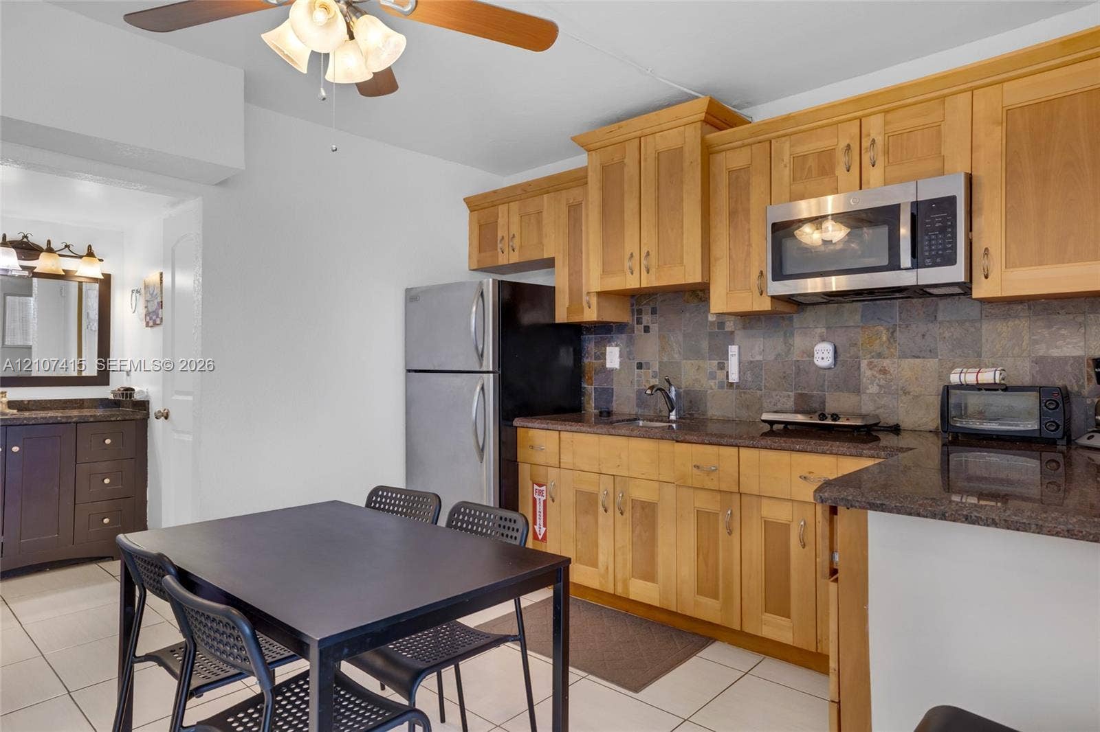 Kitchen featuring natural wood cabinetry, stone-tiled backsplash, stainless steel microwave, stone-surface countertops, and ceiling-mounted light fixture with fan