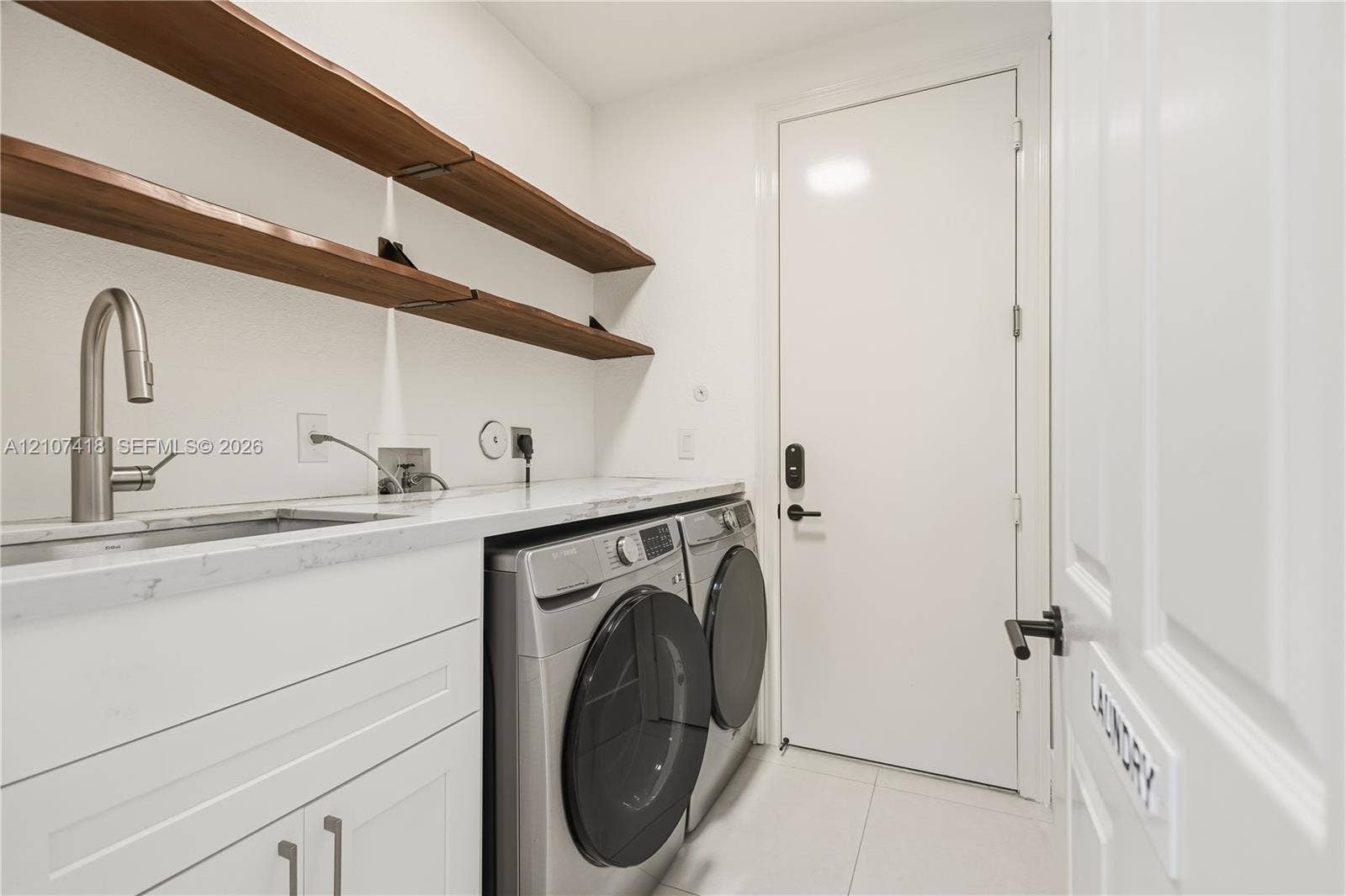 19305 Skyridge Circle, #n/a, Boca Raton, Boca Raton, FL 33498 Laundry room featuring white cabinetry, stone-finish countertops, an undermount sink with a high-arch faucet, wall-mounted wood-finish shelving, and large-format tile flooring