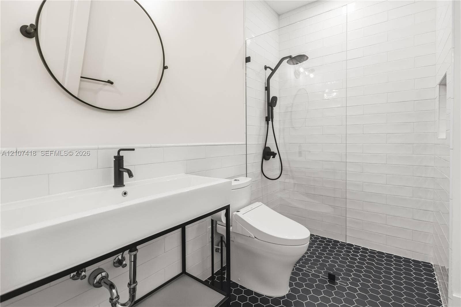 19305 Skyridge Circle, #n/a, Boca Raton, Boca Raton, FL 33498 Wall-mounted round mirror, console-style vanity with a rectangular sink, matte black faucet hardware, glass-enclosed shower with subway tile surround, and hexagonal floor tiling