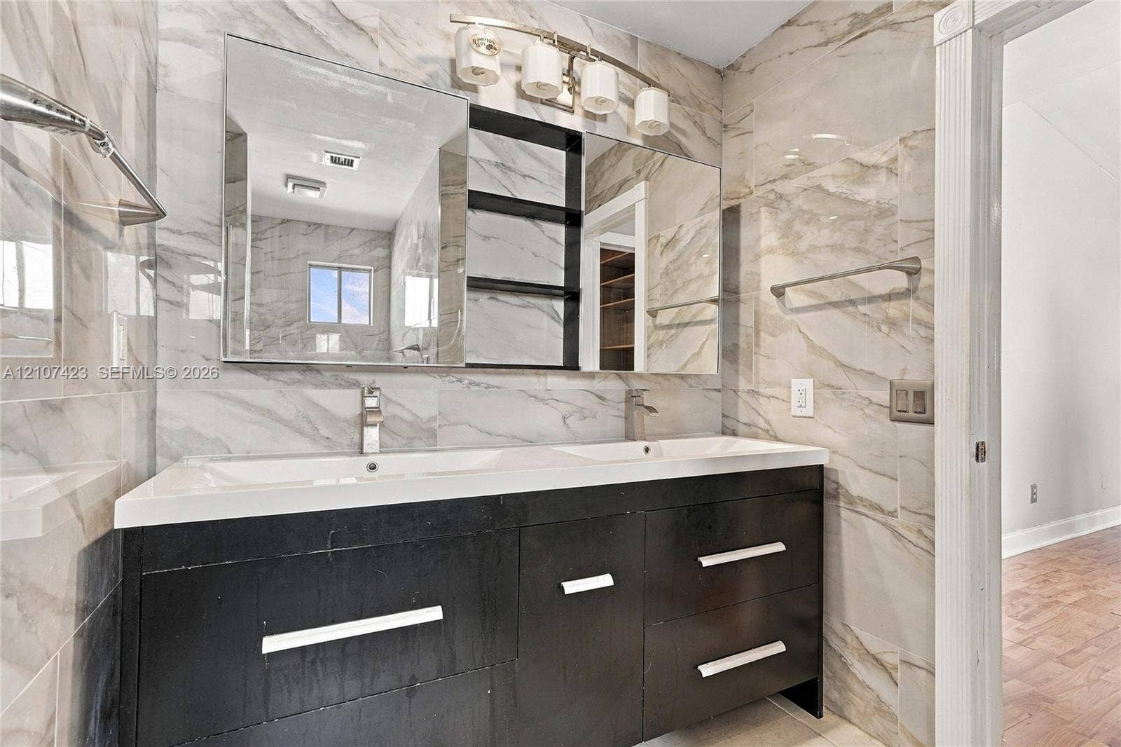 650 NE 195th Street, Miami, FL 33179 Double vanity featuring a dark-toned cabinet base, integrated dual sinks, large-scale tile wall surfacing, polished metal hardware, and multi-light vanity fixtures