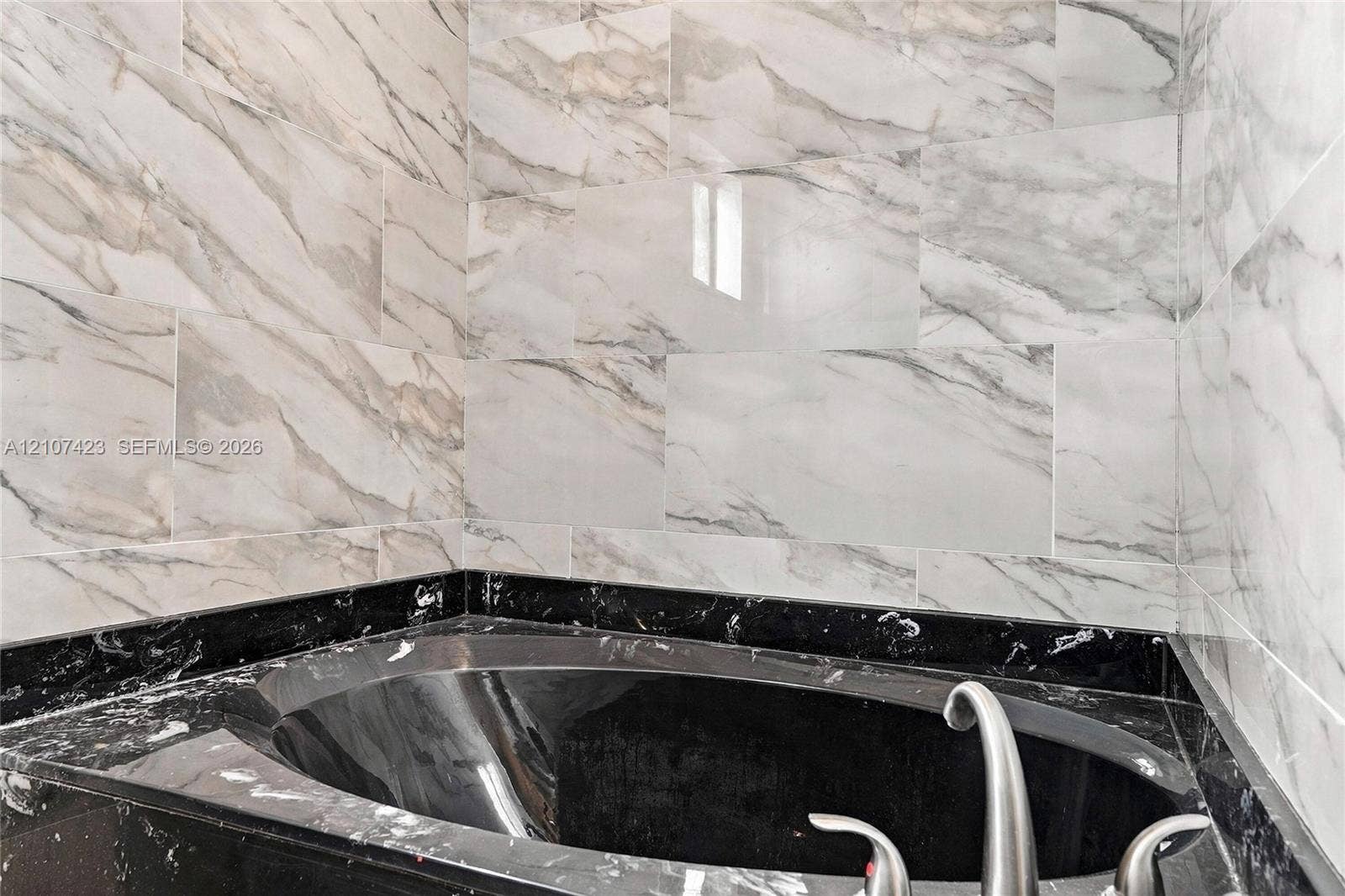 650 NE 195th Street, Miami, FL 33179 Built-in soaking tub with a dark stone-finish surround, complemented by floor-to-ceiling wall tile featuring a veined pattern and a metallic faucet fixture
