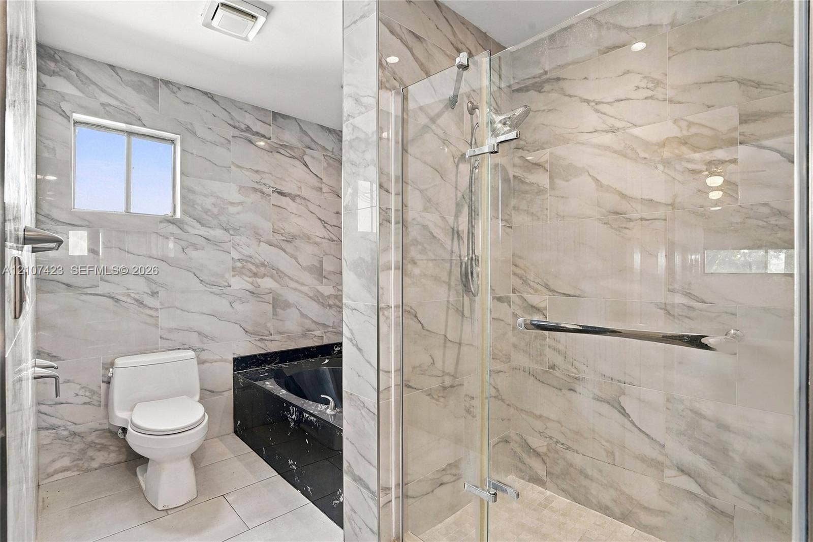 650 NE 195th Street, Miami, FL 33179 Bathroom featuring floor-to-ceiling veined tile, a glass-enclosed shower, a black stone-finish bathtub surround, and a white porcelain toilet