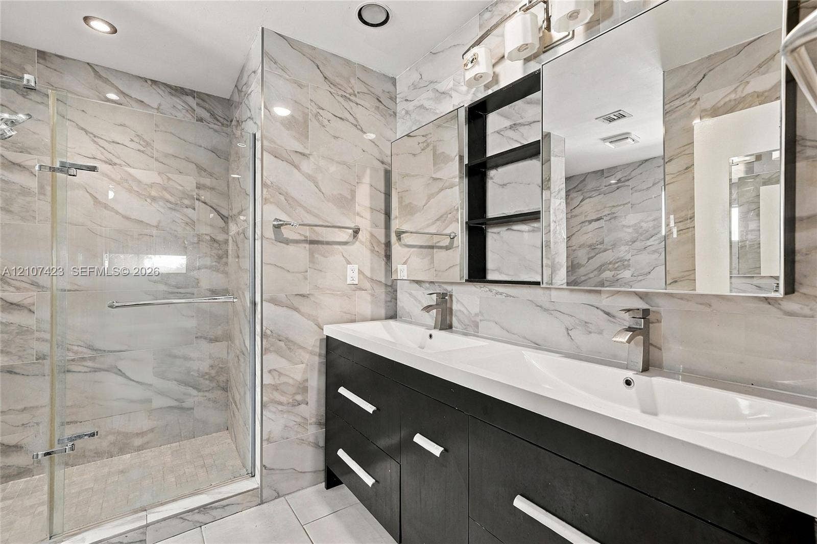 650 NE 195th Street, Miami, FL 33179 Dual-sink vanity with dark cabinetry and metallic hardware, large wall-to-wall mirror, glass-enclosed shower, and full-height wall tile