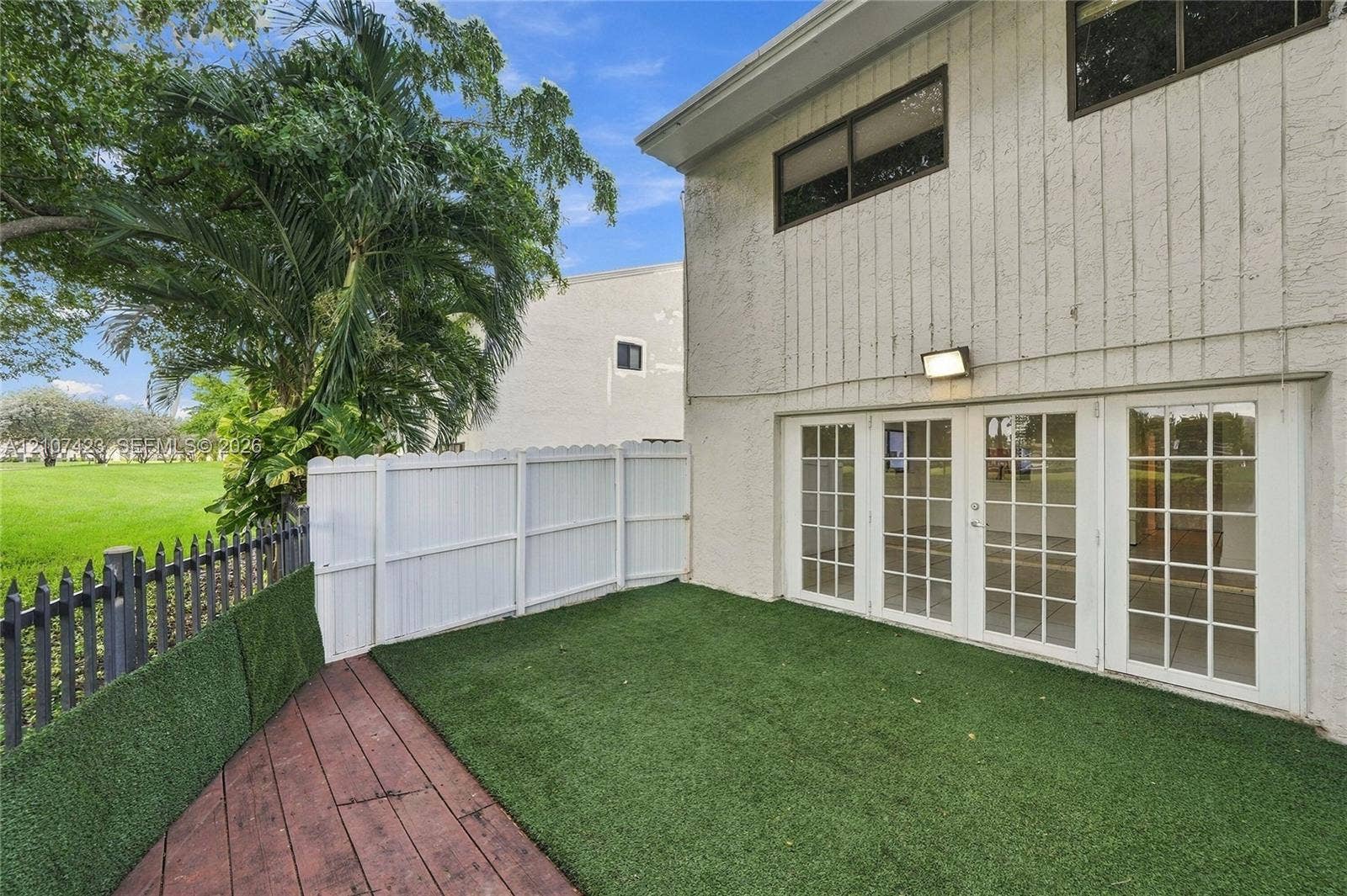650 NE 195th Street, Miami, FL 33179 Exterior featuring vertical siding, four-panel glass doors, synthetic turf lawn, wood-finish decking, and partial privacy fencing