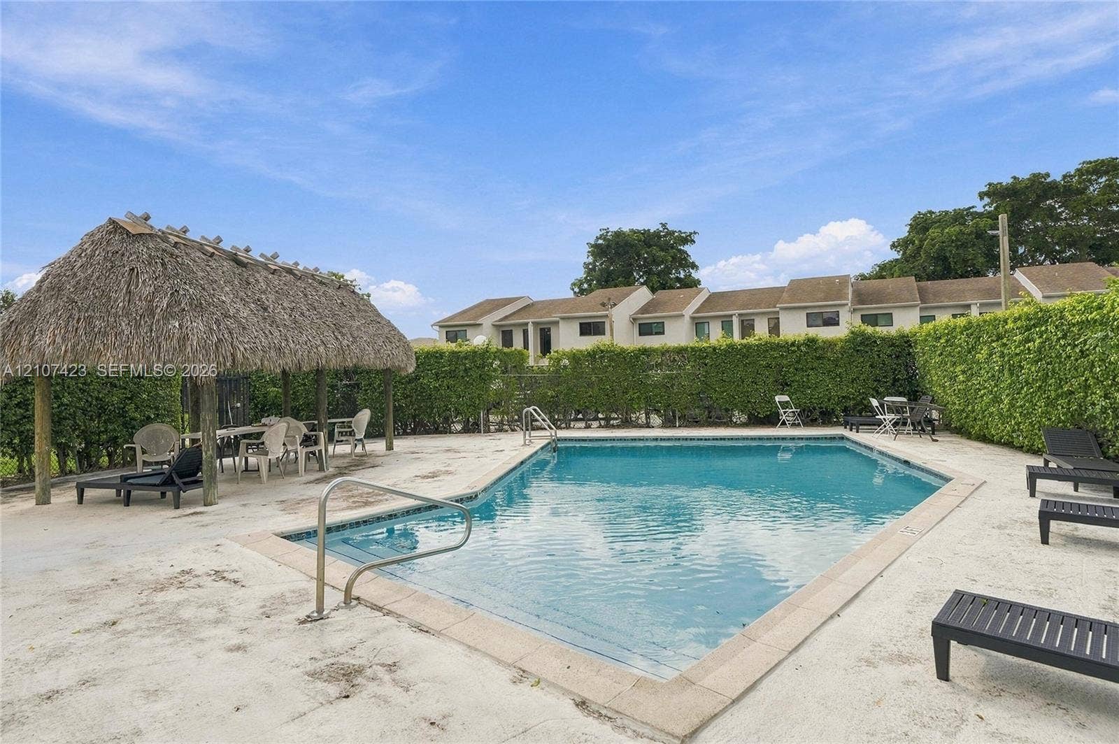 650 NE 195th Street, Miami, FL 33179 Swimming pool with a metal handrail, thatched-roof cabana, and perimeter privacy hedging