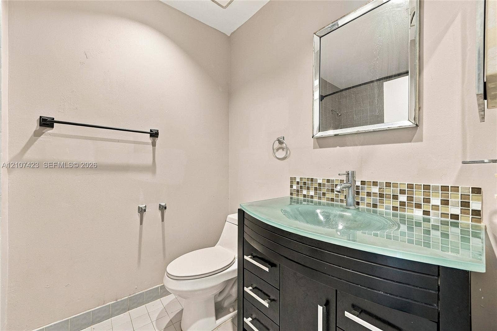 650 NE 195th Street, Miami, FL 33179 Vanity featuring a dark wood-finish cabinet, glass countertop with integrated basin, and mosaic tile backsplash, accented by a chrome faucet and framed mirror