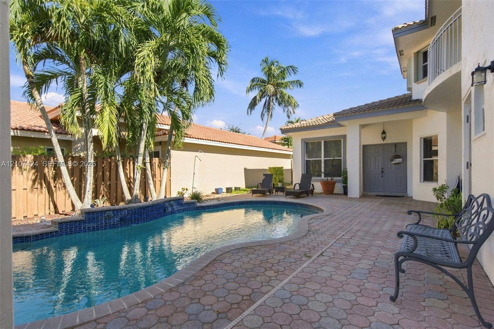 14702 Vista Luna Drive, Davie, FL 33325 Rear exterior featuring a freeform swimming pool with blue tile accents, expansive paver patio, wood privacy fencing, and mature palm trees