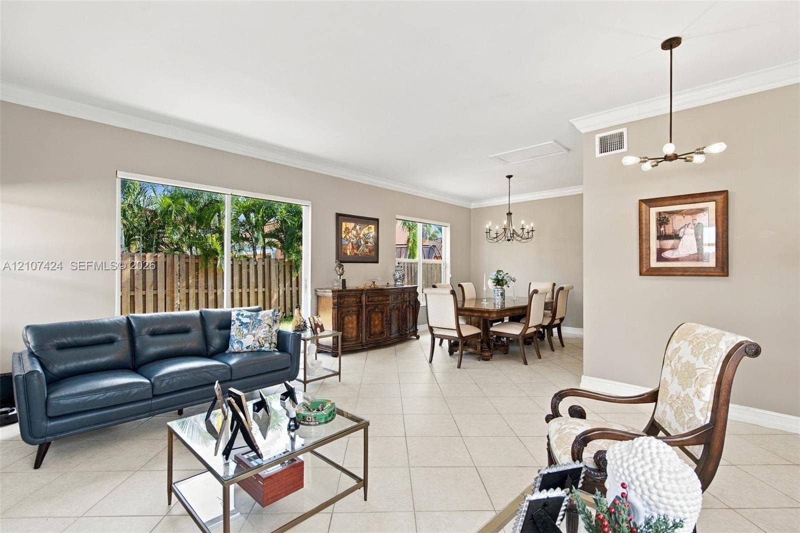 14702 Vista Luna Drive, Davie, FL 33325 Open-concept living space featuring large-format ceramic tile flooring, neutral wall tones, crown molding, sliding glass doors, and two multi-arm chandeliers