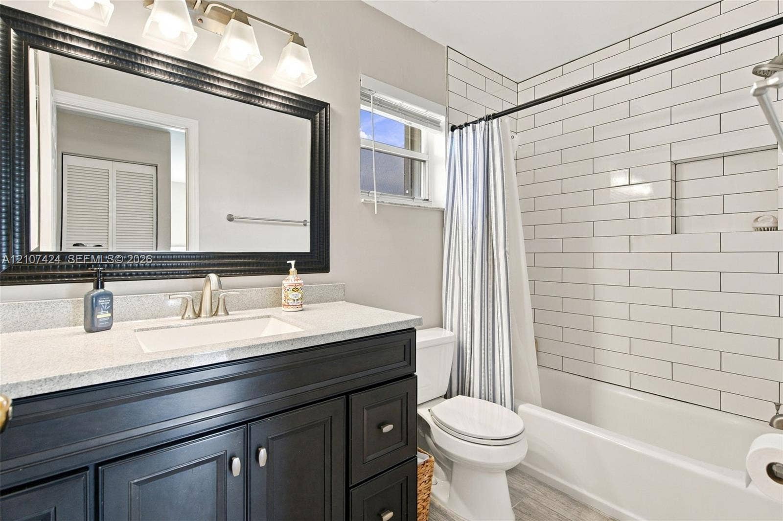 14702 Vista Luna Drive, Davie, FL 33325 Bathroom featuring a dark-toned vanity with a light stone-surface countertop, rectangular undermount sink, framed wall mirror with integrated multi-bulb lighting, and a shower-tub combination finished with horizontal subway tiling and a recessed niche