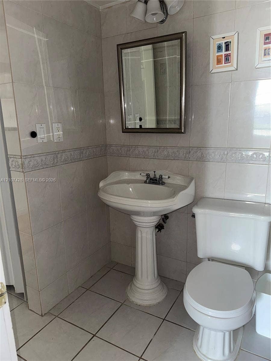 3475 N Country Club Drive, #704, Aventura, Aventura, FL 33180 Full-wall tile surround with decorative accent border, white pedestal sink, polished chrome plumbing fixtures, rectangular wall-mounted mirror, and tile flooring
