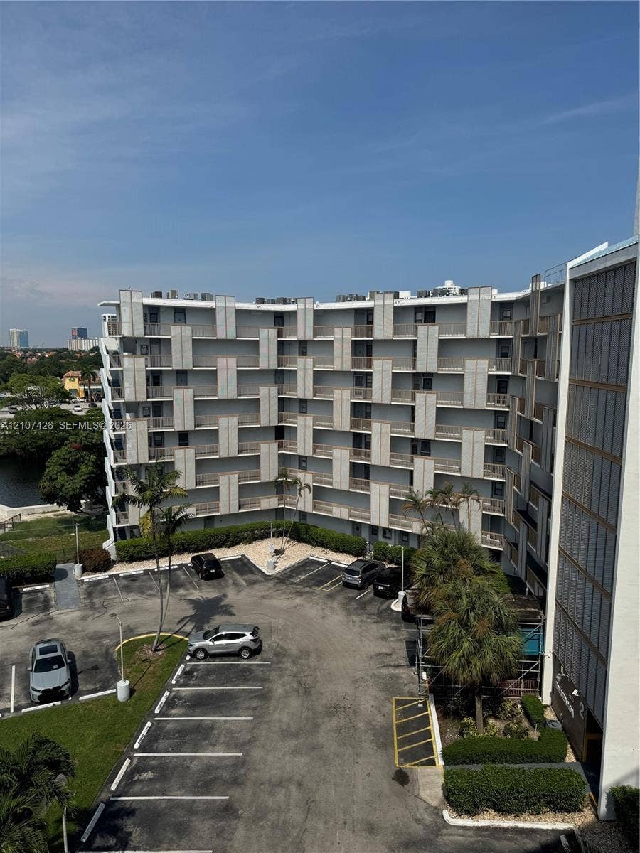 3475 N Country Club Drive, #704, Aventura, Aventura, FL 33180 Mid-rise residential building featuring a structural geometric facade, stacked private balconies, dedicated surface parking, and mature palm landscaping