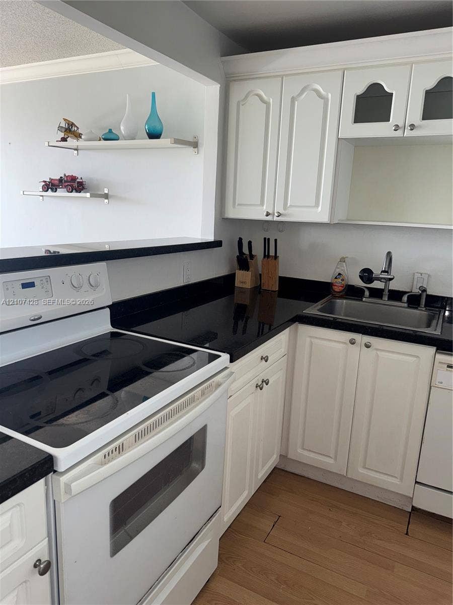 3475 N Country Club Drive, #704, Aventura, Aventura, FL 33180 Kitchen featuring white cabinetry with glass-panel accents, dark countertops, wood-finish flooring, two wall-mounted floating shelves, and an electric range
