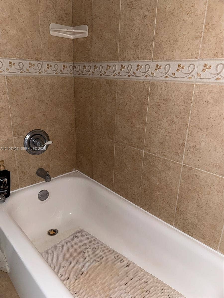 3475 N Country Club Drive, #704, Aventura, Aventura, FL 33180 Bathtub featuring wall-mounted chrome fixtures and full-height stone-finish tile surround with an integrated decorative border and built-in corner shelf