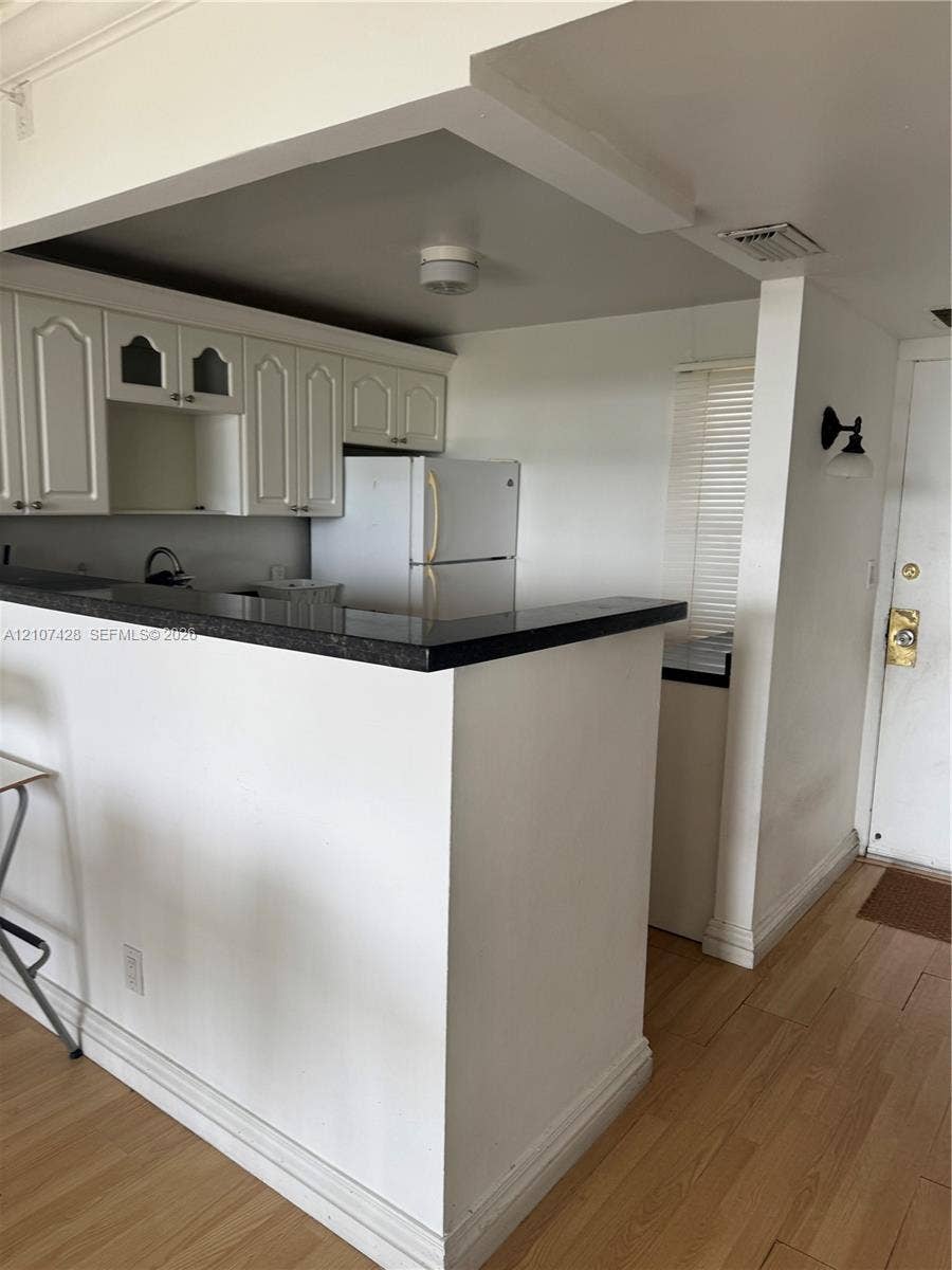 3475 N Country Club Drive, #704, Aventura, Aventura, FL 33180 Kitchen featuring a peninsula with dark stone-finish countertop, white cabinetry with glass-front detailing, wood-finish flooring, and a wall-mounted light fixture