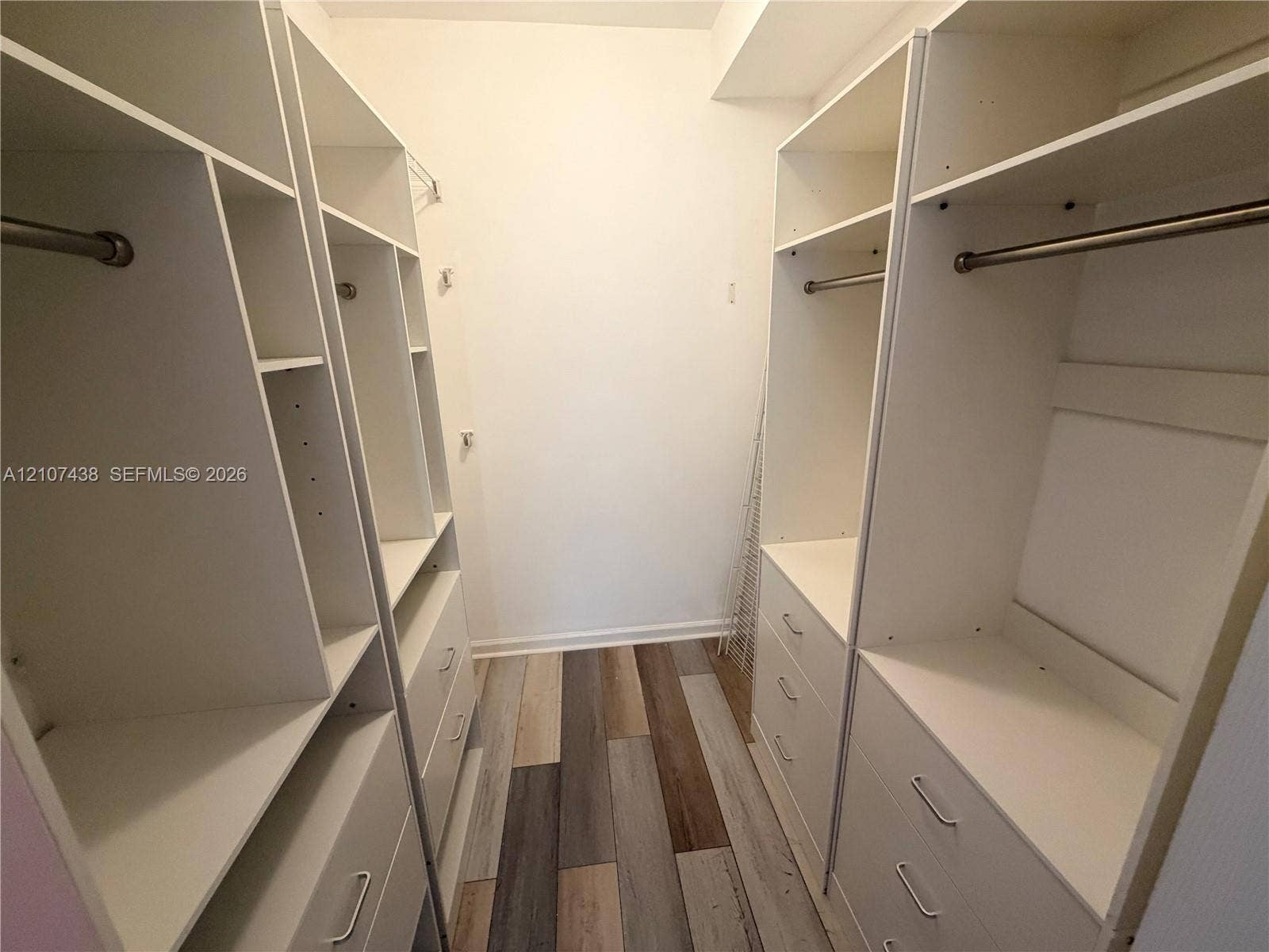 Walk-in closet featuring floor-to-ceiling white built-in shelving, integrated dresser drawers, metal garment rods, and wood-finish flooring